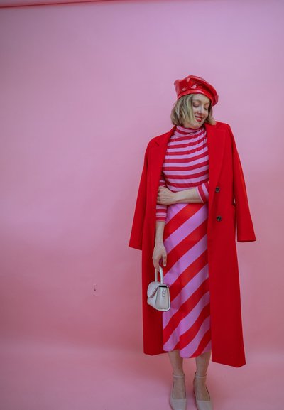 Mujer con abrigo rojo y boina sobre un vestido de rayas rosas y rojas, sosteniendo un pequeño bolso blanco contra un fondo rosa.
