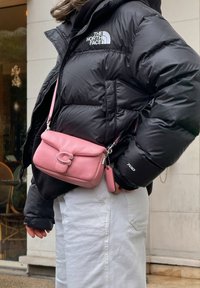 Doudoune noire avec logo, associée à un sac en cuir rose pâle en bandoulière, avec un fermoir et un détail porte-clés, portée sur un pantalon de couleur claire.