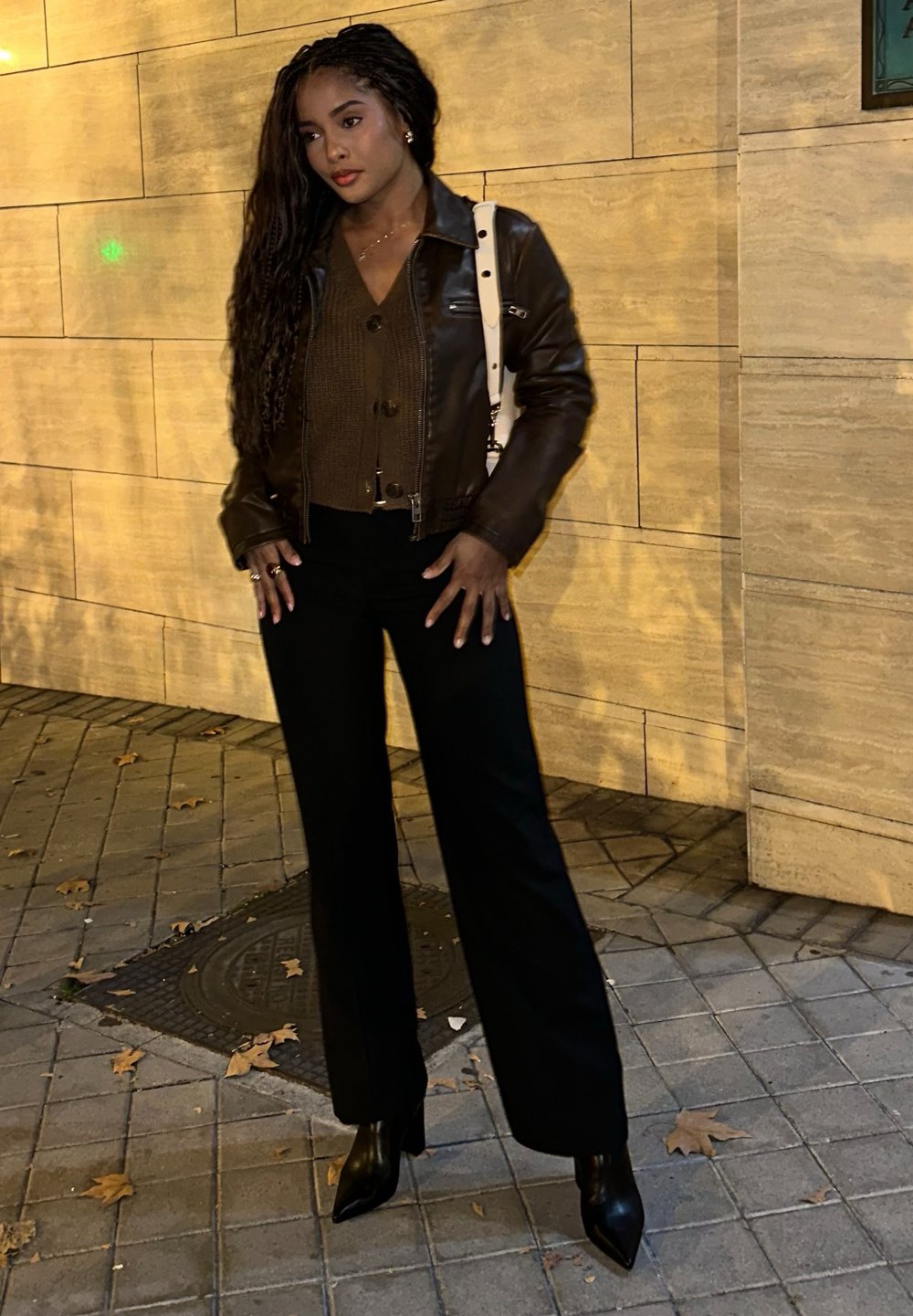 Giacca di pelle marrone sopra un top a maglia marrone scuro, abbinata a pantaloni larghi neri e stivali alla caviglia neri. Sfondo con muro di pietra e pavimentazione.