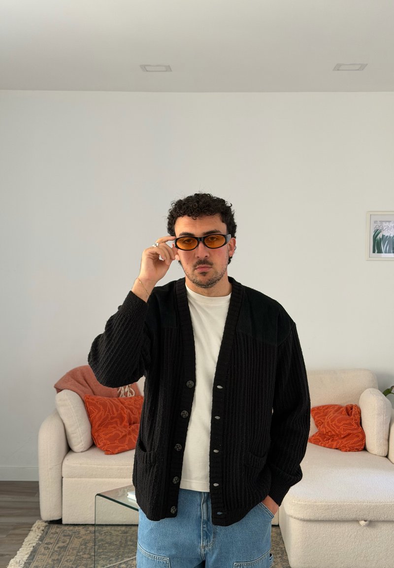 Homem a usar óculos com lentes alaranjadas, cardigan preto, camisa branca e jeans azuis, está em pé numa sala de estar com sofás bege e almofadas alaranjadas.
