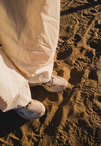 Baskets beige clair avec un matériau texturé, des lacets noirs et des semelles arrondies. Un pantalon ample en tissu crème effleure presque le sol sablonneux.
