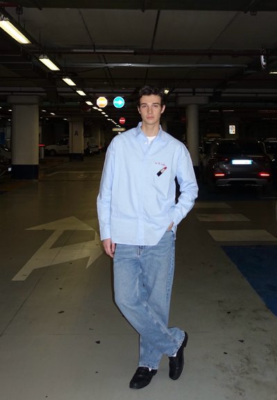 Camisa de botones azul claro con un diseño bordado de lápiz labial, combinada con unos vaqueros holgados azules y zapatos negros, ambientada en un garaje de aparcamiento con poca luz.