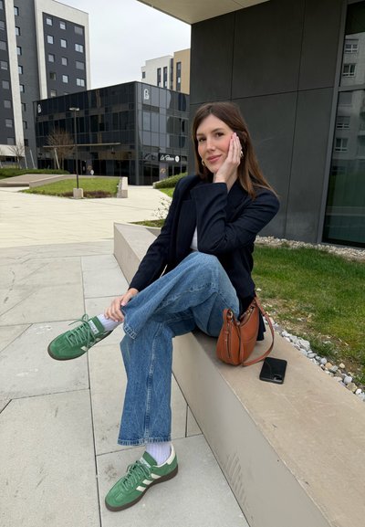 Joven sentada en un borde de piedra, vestida con un blazer azul marino, una blusa blanca y jeans de mezclilla claros. Zapatillas verdes con rayas blancas, bolso marrón cerca.