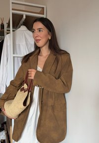 Hnědý velký semišový blejzr s knoflíkovým zapínáním a dvěma předními kapsami. Pod tím je bílý krajkový detail. Model drží tkanou slaměnou kabelku.