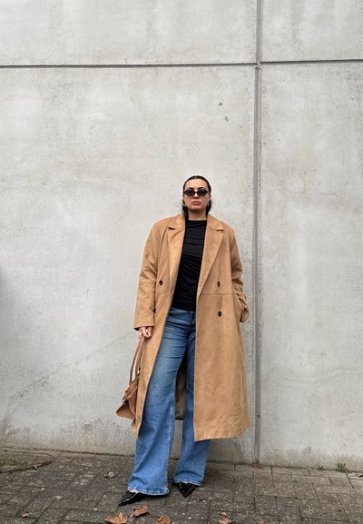 Mujer vistiendo un abrigo largo color beige, camiseta negra, vaqueros azules, zapatos negros puntiagudos, gafas de sol y sosteniendo un bolso a juego color beige, de pie junto a una pared de hormigón.