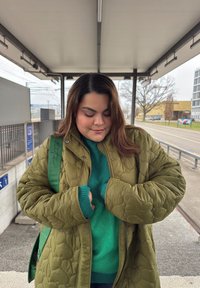 Oliivinvihreä tikkitakki, jossa on suuret ompeleet, päällä kirkkaanvihreä neule. Takissa on pitkät hihat ja sivussa vihreä reppu.