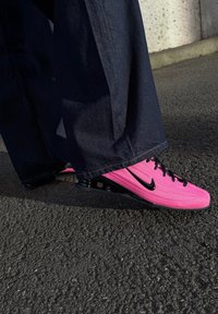 Baskets Nike rose vif avec un swoosh noir et des lacets noirs, portées avec un jean large foncé sur une surface d'asphalte texturée.