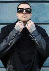 Poncho en mélange de laine noire avec accents en cuir, col montant et manches à zip. Comprend plusieurs anneaux et de grandes lunettes de soleil noires.