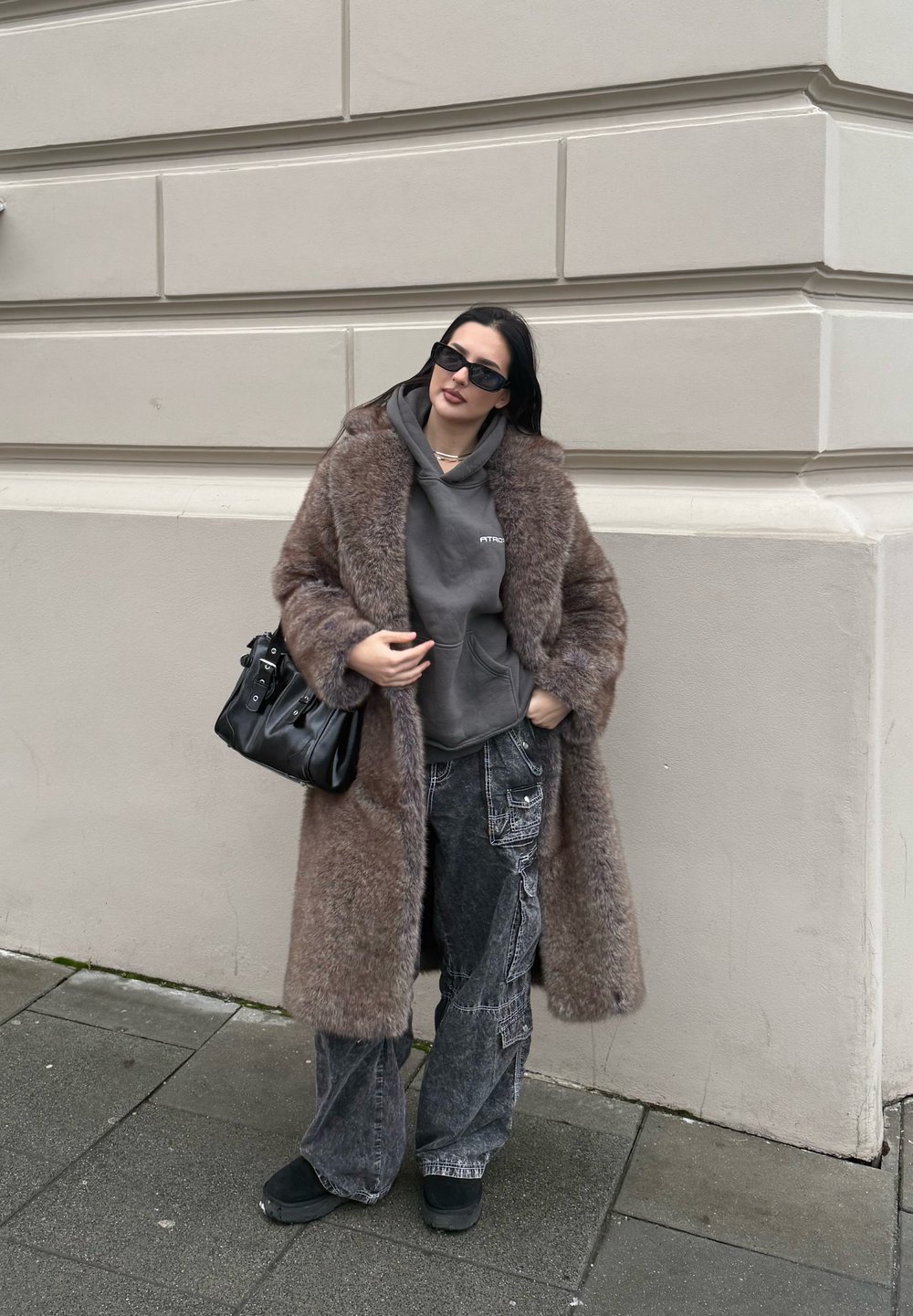 Μακρύ καφέ ψεύτικο γούνινο παλτό, γκρίζα κουκούλα, φθαρμένα μαύρα φαρδιά τζιν, μαύρα γλυκά παπούτσια και υπερμεγέθη μαύρα γυαλιά ηλίου. Παρουσιάζονται υφές υλικών.