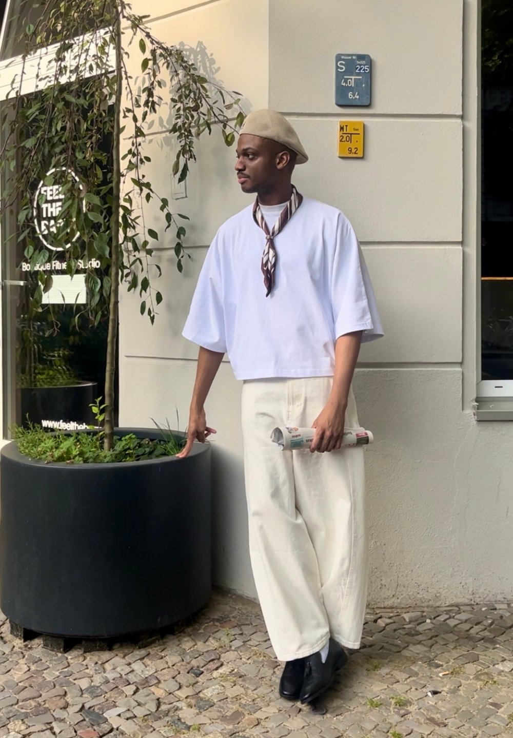 Vit croppad t-shirt, beige beret, brun mönstrad halsduk, krämfärgade vida byxor och svarta skor. Håller en rullad tidning, står vid en växt.