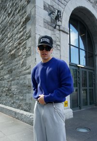 Blå strikket sweater, grå vide bukser med kædetræk, sort kasket med "DC Shoes" logo, mørke solbriller, urban sten baggrund.
