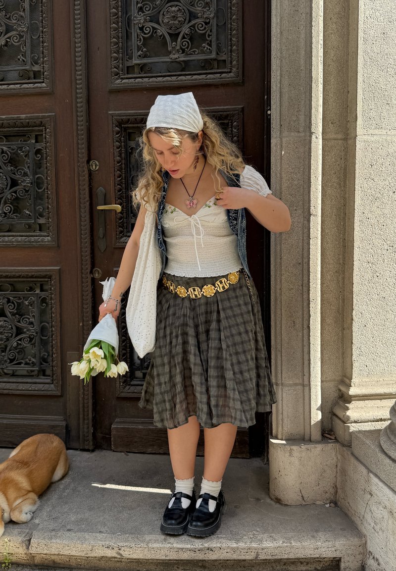 Jeune femme en jupe à motifs et top blanc, tenant des tulipes blanches, debout sur une marche en pierre près d'une porte en bois ornée, avec un petit chien couché à proximité.