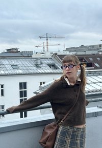 Pulover maro tricotat cu guler și nasturi, asortat cu o fustă în carouri. O geantă maro tip crossbody și ochelari rotunzi completează ținuta.