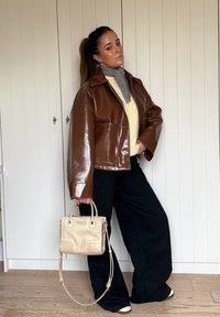 Brown leather cropped jacket, grey jumper, black wide-leg trousers, holding a beige handbag with a smooth texture and a minimalistic design.