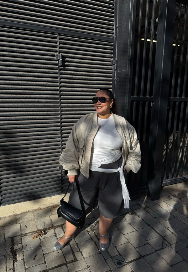 Una mujer con gafas de sol, una chaqueta clara y una falda oscura está de pie en una acera pavimentada, con un bolso negro junto a una verja metálica.