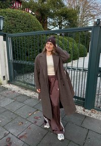 Brun ullfrakk over en beige topp, kombinert med skinnende brune treningsbukser med hvite striper. Brun strikket lue og grå joggesko fullfører antrekket.