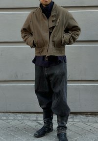 Jachetă verde măsliniu cu un guler texturat, purtată peste o cămașă închisă la culoare. Pantaloni negri lejeri și cizme negre până la vițel completează ținuta.