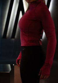 Fitted maroon long-sleeved top with a high collar, smooth texture, and subtle pattern; paired with black sports leggings featuring a logo.