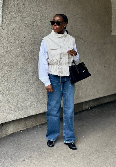 Mujer con gafas de sol, chaleco blanco sobre camisa blanca extragrande, vaqueros azules, zapatos negros, sosteniendo bolso negro, de pie contra una pared texturizada.