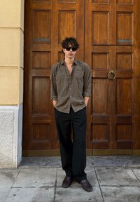 Donkere olijfgroen overhemd met opgerolde mouwen, zwarte broek en donkere schoenen, staande tegen een houten deur met bronzen beslag.