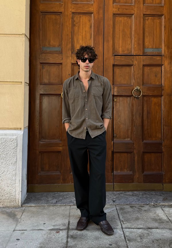 Chemise button-up vert olive foncé avec des manches retroussées, pantalon noir et chaussures foncées, se tenant devant une porte en bois avec des quincailleries en bronze.