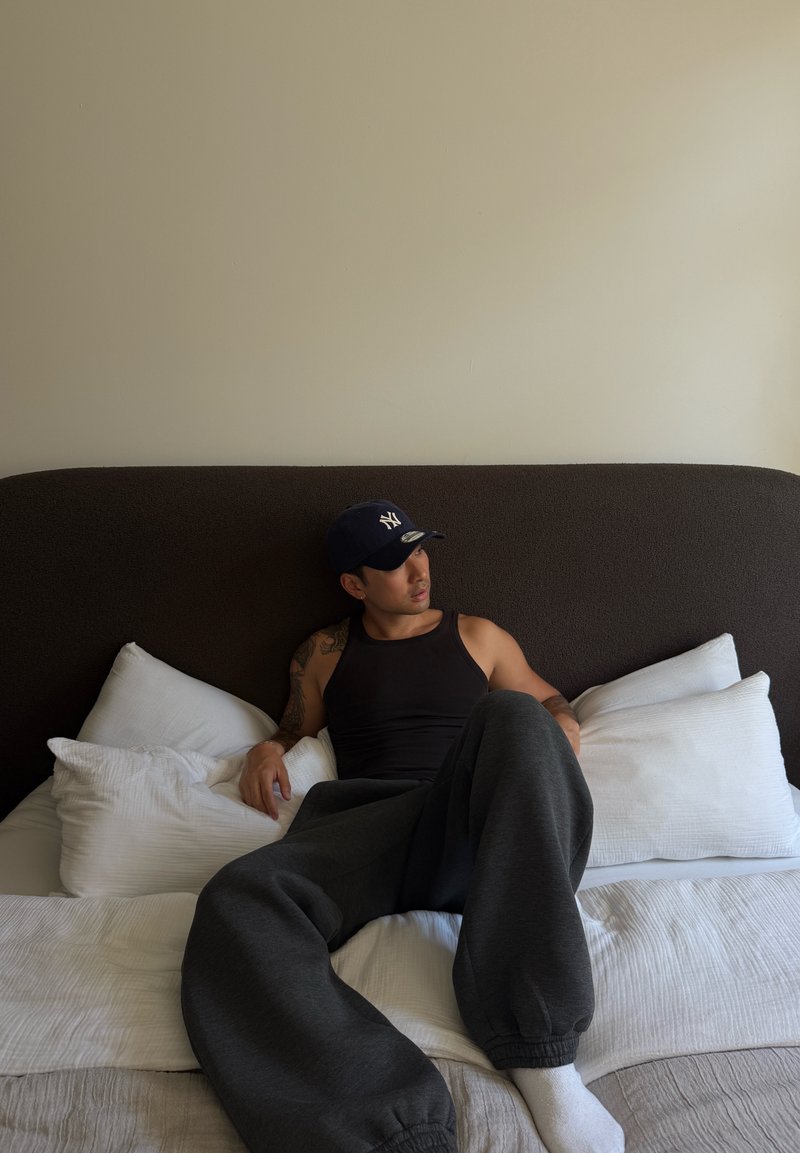 Homme portant un débardeur noir, un pantalon de survêtement sombre et une casquette de baseball assis sur un lit avec des oreillers blancs et un mur beige en arrière-plan.