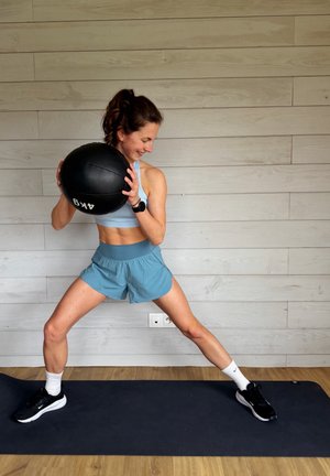 Mujer con ropa deportiva sostiene una pelota medicinal de 4 kg, de pie sobre una colchoneta con las piernas separadas en una posición de zancada lateral en interiores.