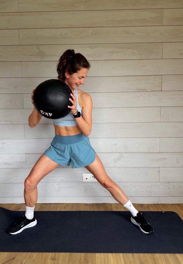 Kvinde i sportstøj holder en medicinbold på 4 kg, stående på en måtte med benene spredt i en sidelungeposition indendørs.