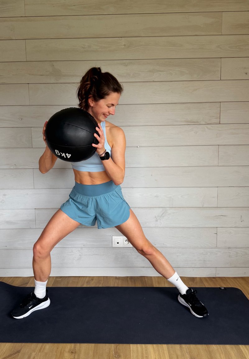 Γυναίκα με αθλητική ένδυση κρατά μια ιατρική μπάλα βάρους 4 κιλών, στέκεται σε στρώμα με τα πόδια ανοιχτά σε θέση πλευρικής προβολής σε εσωτερικό χώρο.