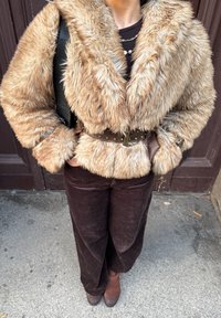 Fuskepelsjakke i lys brun med bred krage, belte med brunt naglebelte. Kombinert med mørkebrune kordfløyelsvide bukser og brune støvler.