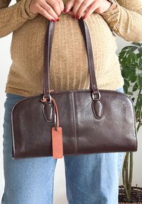 Personne avec du vernis à ongles rouge tenant un sac à main en cuir marron foncé avec des longues lanières et une étiquette orange attachée.