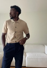 Rövid göndör hajú férfi, bézs színű kötött rövid ujjú ingben, tengerészkék nadrágban, ezüst gyűrűvel, órával és lógó fülbevalóval, egy fehér kanapé mellett áll.