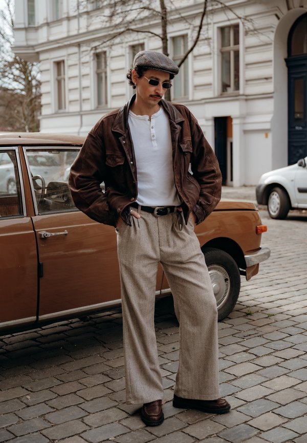 Mann i brun skinnjakke, beige bukser, flat caps og solbriller som står på brosteinsgate ved en brun veteranbil.