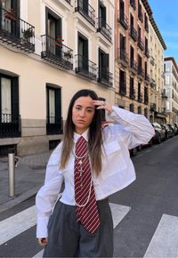 Camisa curta branca, gravata listrada em vermelho e branco, colares de pérolas em camadas. Calças largas cinza. Ambiente urbano com edifícios e carros estacionados.