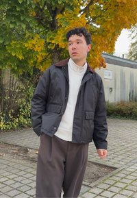 A young man dressed in a black jacket and a white zip-up sweater stands on a paved area beneath a tree adorned with green and yellow autumn leaves.