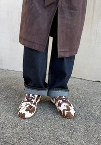 Barna és fehér mintás, velúrból készült sportcipő, fűzős kialakítással, az alsó maradék részén felgombolt sötét farmernadrággal, amely kontrasztban áll egy barna kabáttal.