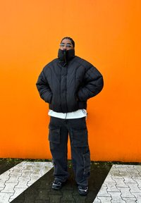 Crna puf jakna s visokim kragnom, nosi se preko bijele košulje. Tamni cargo hlače s džepovima, i crne tenisice, postavljene nasuprot narančastoj zidu.