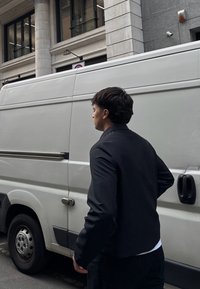 Giacca nera dal taglio aderente, indossata sopra una camicia bianca, abbinata a pantaloni scuri, vista da dietro, accanto a un furgone bianco per le consegne.