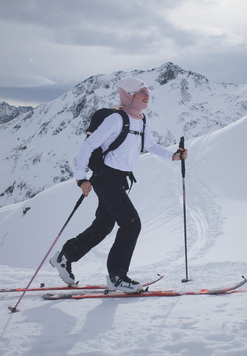 Personne faisant du ski en montée sur une montagne enneigée avec des bâtons, portant une veste blanche, un pantalon noir, un couvre-visage rose et un sac à dos sous un ciel nuageux.