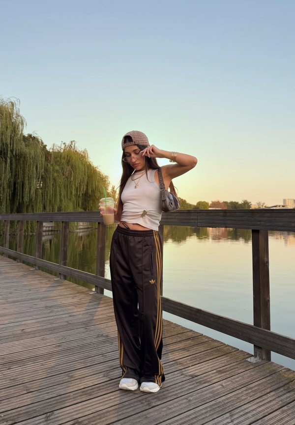 Jonge vrouw staat op een houten vlonder bij het meer bij zonsondergang, draagt een pet, wit tanktop, zwarte joggingbroek, houdt een ijskoud drankje vast.