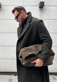 Homme portant un pardessus vert foncé et des lunettes de soleil, tenant un grand sac en suède marron plié, debout contre un mur carrelé gris.