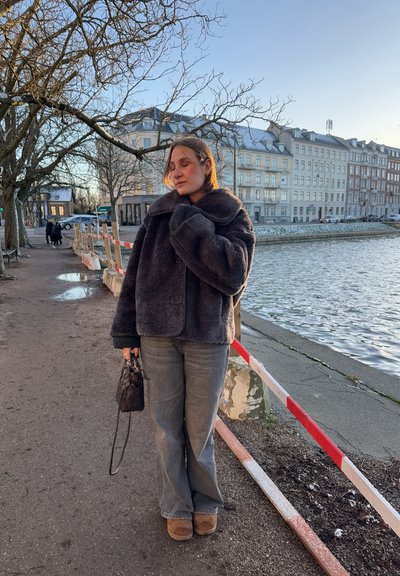 Chaqueta de piel sintética gris con un gran cuello, combinada con jeans grises holgados y botas marrones. Sosteniendo un pequeño bolso negro junto al agua.