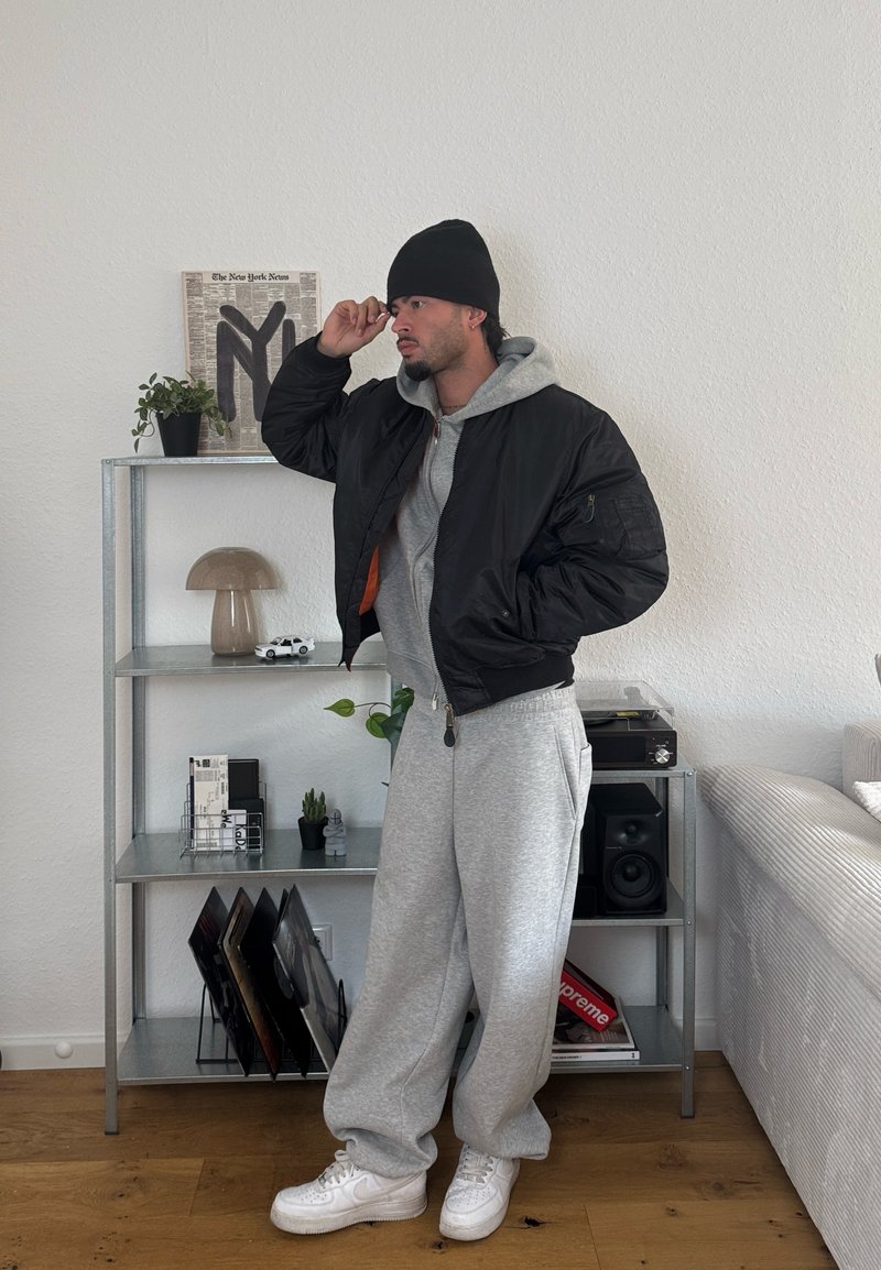 Moški z črnim klobukom, črnim jopičem, sivo kapuco in trenirkami stoji notranjosti ob kovinskih policah z okrasnimi predmeti in kavčem.