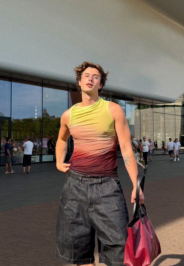 Jonge man draagt een mouwloze top met kleurverloop en oversized donkere shorts, houdt een rode handtas vast en staat buiten een modern glazen gebouw.