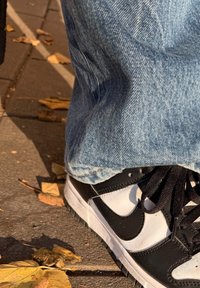 Zapatillas Nike en blanco y negro con cuero texturizado, cordones negros y suela de goma, combinadas con unos vaqueros de mezclilla azul claro, descansando sobre una acera con hojas.