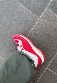 Κόκκινο και λευκό αθλητικό παπούτσι με υφασμάτινη επάνω επιφάνεια, ορατό λογότυπο, στρωματοποιημένο σχέδιο και καουτσούκ σόλα, φορεμένο με καφέ παντελόνι πάνω σε πέτρινο δάπεδο.