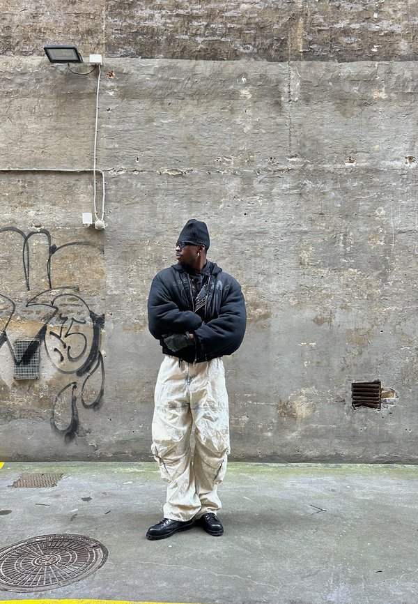Black oversized puffer jacket, off-white cargo pants, black boots, textured wall background, with graffiti and exposed electrical fixtures.