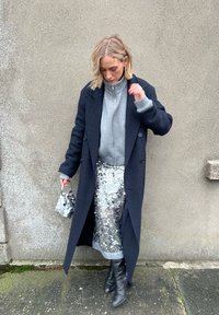 Femme debout contre un mur texturé, portant un long manteau sombre, un pull gris à fermeture éclair, une jupe argentée à sequins, des bottes noires et tenant un petit sac.