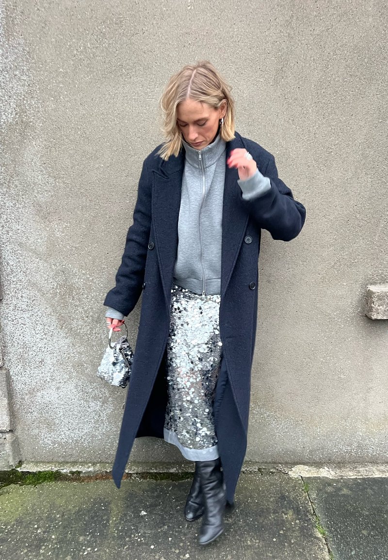 Woman standing against a textured wall wearing a dark long coat, grey zip-up sweater, silver sequin skirt, black boots, and holding a small bag.