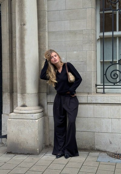 Jersey negro de punto y pantalones holgados negros, combinados con un bolso de estampado a cuadros. De pie contra una pared de piedra con una ventana decorativa.